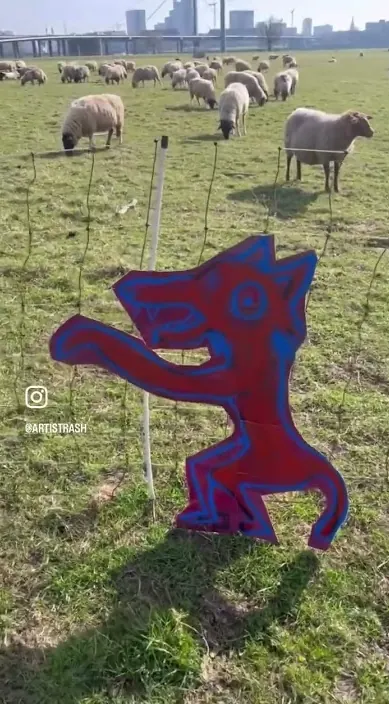 Gemalte Wolfsfigur auf einer Tafel auf einem Feld zwischen Schafen, Art Is Trash